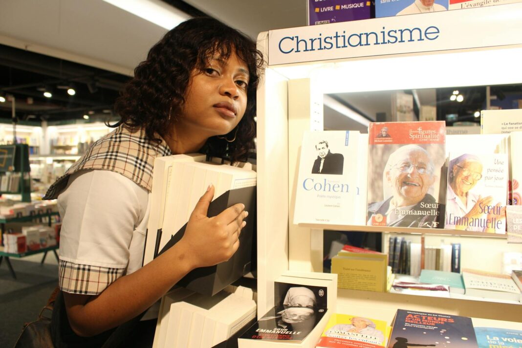 Black woman with short hair holding books in a bookstore aisle.