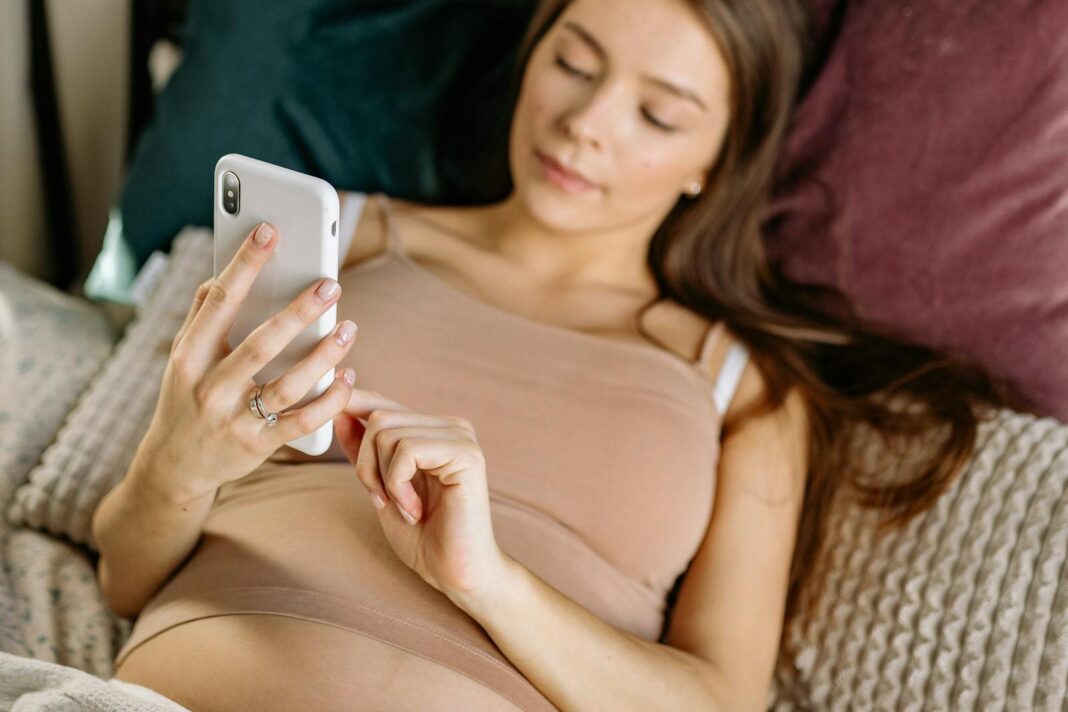 Caucasian pregnant woman lying on bed using smartphone, embodying modern lifestyle.