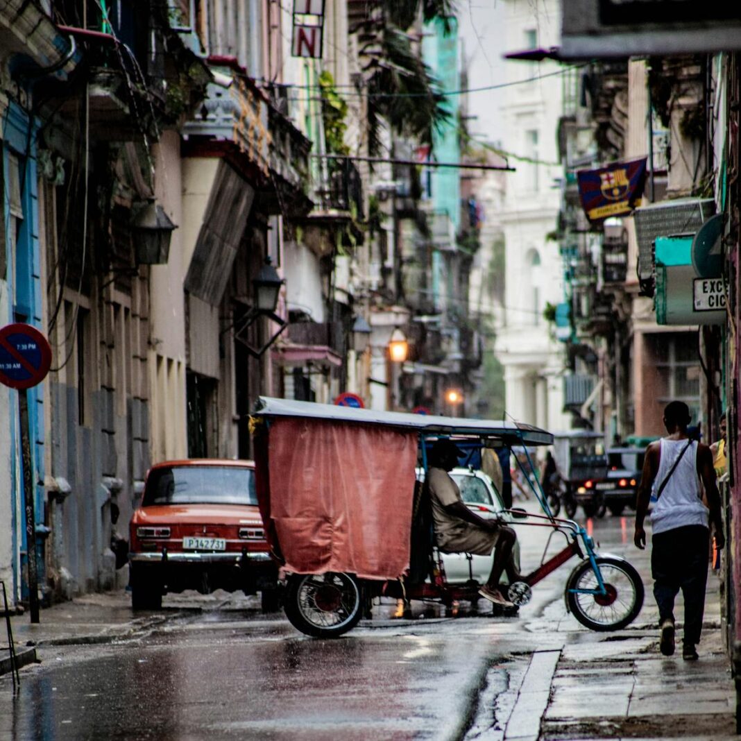 A bustling street in Havana, Cuba features classic cars, a rickshaw, and vibrant architecture.