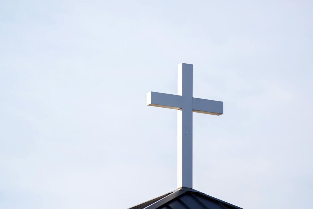 Minimalist image of a white cross on a church roof against a clear blue sky.