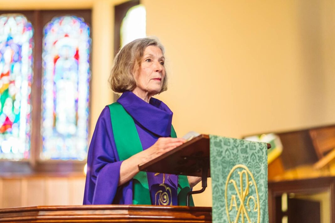 An elderly woman delivers a sermon from a lectern in a beautifully lit church.