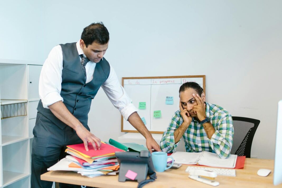 Two businessmen in an office, one stressed at the desk while another points at folders, discussing workload.